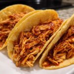 Close-up of three delicious chicken tacos filled with shredded chicken coated in a rich red sauce.