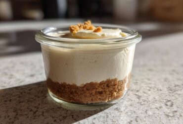 A single serving Protein Cheesecake layered in a small glass jar with a graham cracker crust and crumb topping.