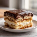 A close-up of a decadent slice of eclair cake with layers of cream, pudding, and chocolate ganache topping.