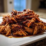 A close-up of tender, saucy Slow cooker pulled pork piled high on a white square plate.