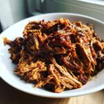 Close-up of tender, shredded Slow cooker pulled pork coated in a rich, dark sauce served in a white bowl.