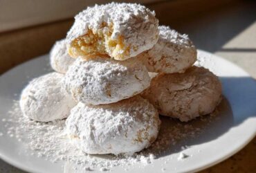 A stack of soft, powdered sugar-dusted cool whip cookies on a white plate, one cookie is broken open.