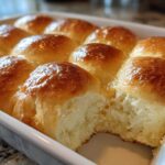 A close-up of golden brown, glossy sourdough discard rolls baked together in a white rectangular dish, with one roll pulled apart showing the soft interior.