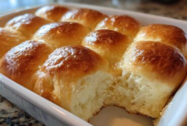 A close-up of golden brown, glossy sourdough discard rolls baked together in a white rectangular dish, with one roll pulled apart showing the soft interior.