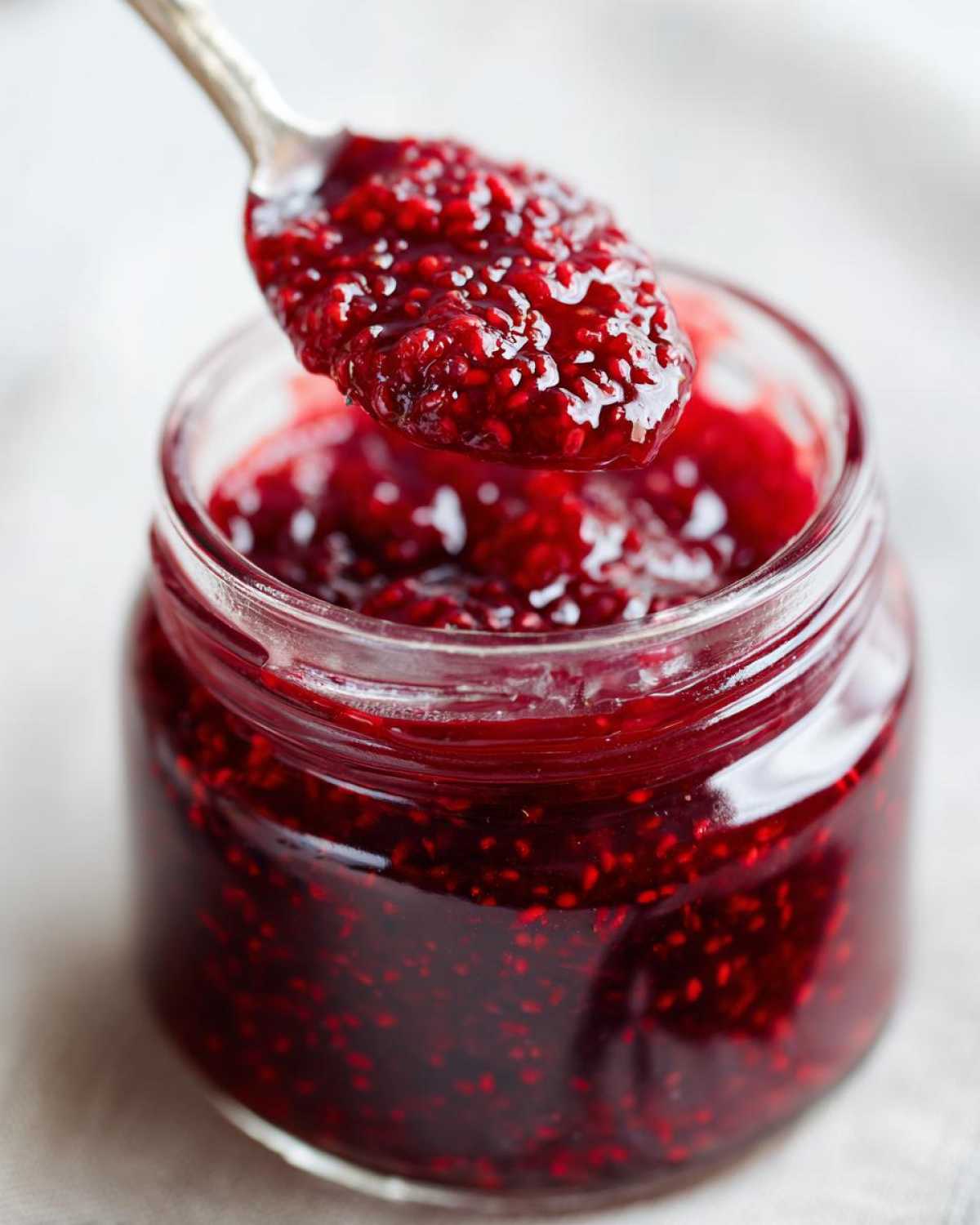 A spoonful of glistening, chunky homemade raspberry jam is lifted from a small glass jar.