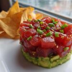 Close-up of stacked tuna tartare over avocado, garnished with green onions and sesame seeds, served with tortilla chips.