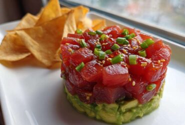 Close-up of stacked tuna tartare over avocado, garnished with green onions and sesame seeds, served with tortilla chips.