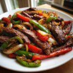 A close-up shot of perfectly cooked steak fajitas featuring sliced steak, vibrant red and green bell peppers, and caramelized onions on a white plate.