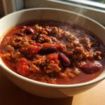 Close-up of a steaming white bowl filled with rich, red high protein chili featuring ground meat and kidney beans.