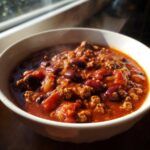 A steaming white bowl filled with rich, hearty high protein chili featuring ground meat and dark red kidney beans.