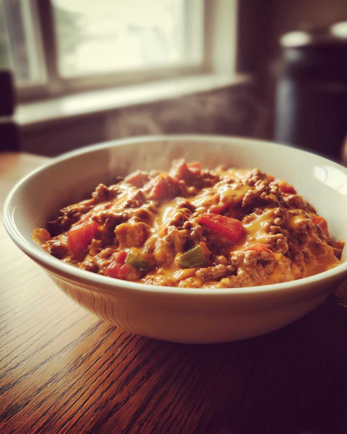 A close-up of a steaming white bowl filled with cheesy, meaty rotel dip featuring ground beef and diced tomatoes.