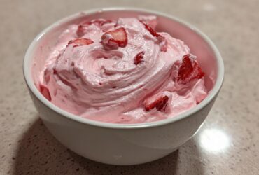 A white bowl filled with fluffy, pink strawberry cheesecake dip topped with fresh strawberry slices.