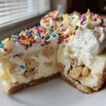 Cross-section view of a slice of sugar cookie cheesecake loaded with cookie chunks, topped with whipped cream and colorful sprinkles.
