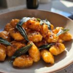 A close-up of bright orange sweet potato gnocchi tossed in sauce and topped with crispy sage leaves.