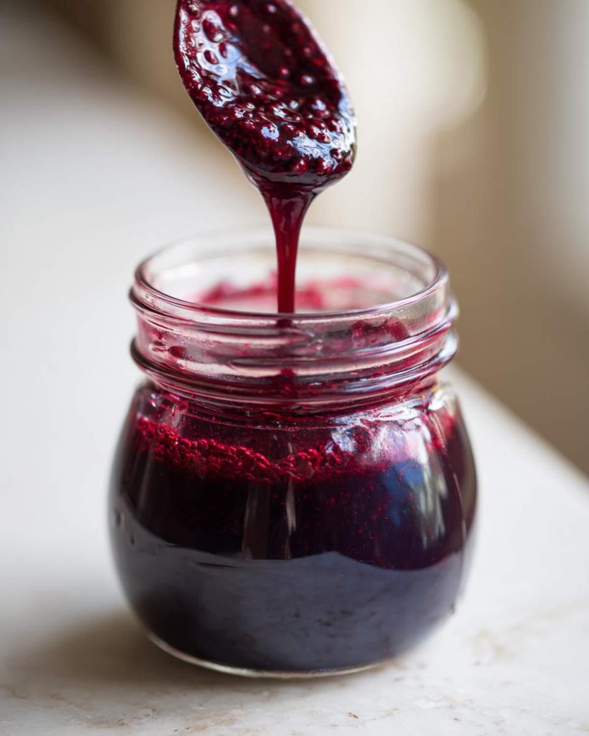 Close-up of thick, dark purple elderberry syrup recipe dripping from a spoon into a small glass jar.