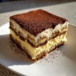 A close-up of a perfect square slice of tiramisu recipe, showing layers of cream, ladyfingers, and a dusting of cocoa powder.