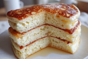 Close-up of a stack of three incredibly fluffy pancakes cut to show the airy interior, drizzled with syrup.