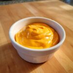 A small white bowl filled with thick, bright orange nacho cheese sauce sitting on a wooden cutting board.