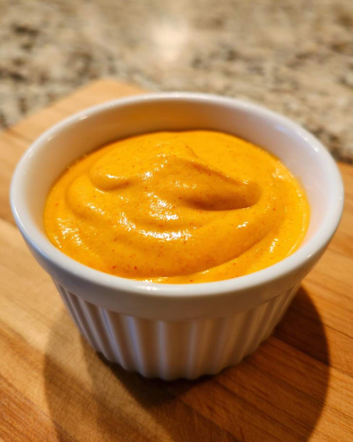 Close-up of a small white ramekin filled with vibrant, smooth nacho cheese sauce, resting on a wooden cutting board.