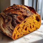 Close-up of a deeply colored, open-crumb pumpkin sourdough loaf cut in half on a marble surface.