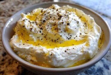 Close-up of a bowl filled with creamy whipped feta dip, drizzled generously with golden olive oil and sprinkled with black pepper.