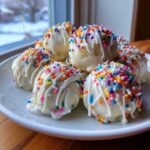 A plate piled high with white chocolate-dipped sugar cookie truffles, drizzled with white icing and covered in rainbow sprinkles.