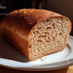 A freshly baked loaf of whole wheat bread, sliced to show the soft, brown crumb, sitting on a white plate.