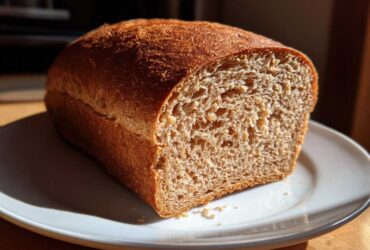 A freshly baked loaf of whole wheat bread, sliced to show the soft, brown crumb, sitting on a white plate.