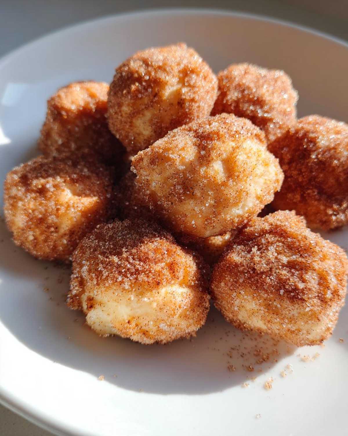 A pile of golden Air Fryer Cinnamon Sugar Churro Bites coated heavily in cinnamon sugar on a white plate.