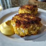Two golden brown Air Fryer Crispy Crab Cakes served on a white plate with a dollop of bright yellow lemon aioli.