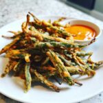 A pile of Air Fryer Crispy Tempura Green Beans served on a white plate with a side of orange dipping sauce.
