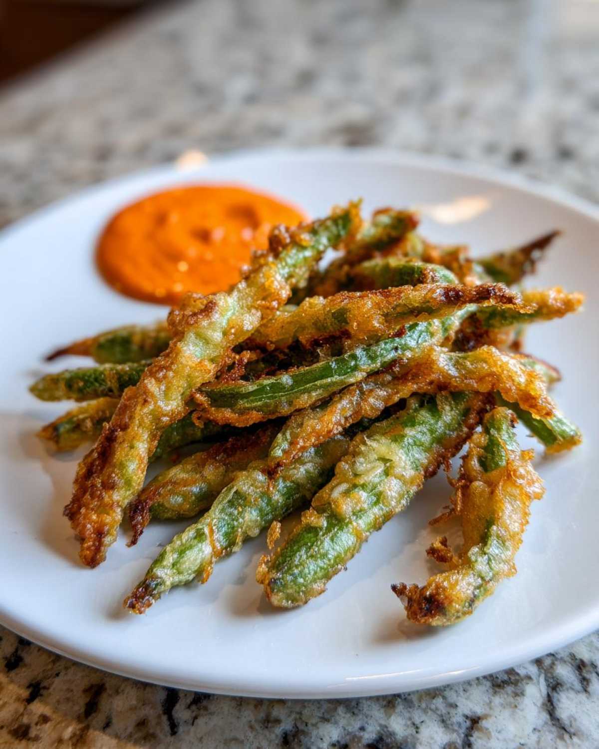 A pile of golden brown Air Fryer Crispy Tempura Green Beans served on a white plate with a side of orange dipping sauce.