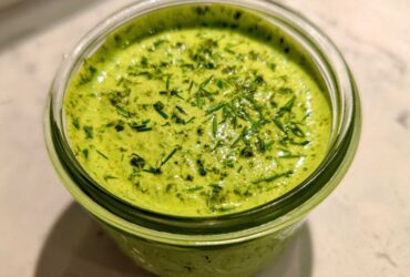 Close-up of vibrant green Copycat Panera Green Goddess Dressing topped with fresh herbs in a glass jar.