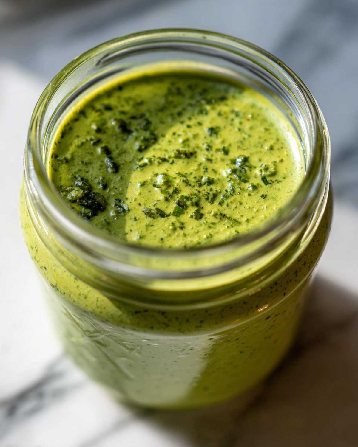 Close-up of vibrant green Copycat Panera Green Goddess Dressing blended smooth in a glass jar, topped with herbs.