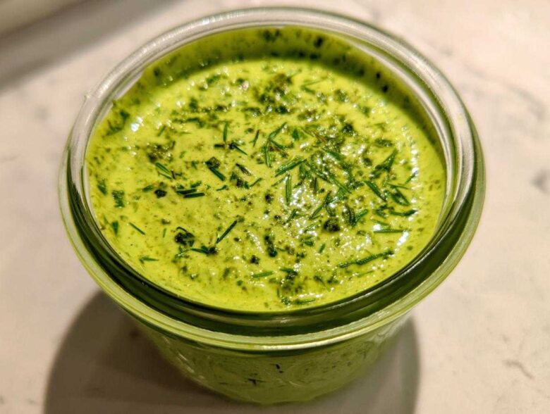 Close-up of vibrant green Copycat Panera Green Goddess Dressing topped with fresh herbs in a glass jar.