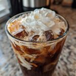 Close-up of a tall glass filled with an iced white chocolate mocha, ice cubes, and topped with whipped cream.