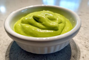 A close-up of vibrant green, thick Creamy Avocado Cilantro Sauce swirled in a small white ramekin.