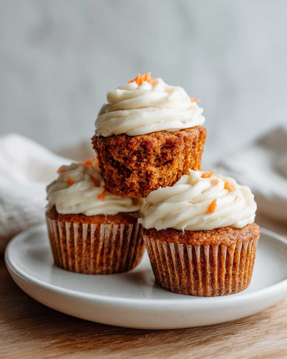 Three Gluten-Free Carrot Cake Cupcakes stacked on a white plate, topped with thick cream cheese frosting.