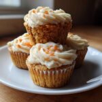Four Gluten-Free Carrot Cake Cupcakes stacked on a white plate, topped with thick cream cheese frosting and shredded carrot.