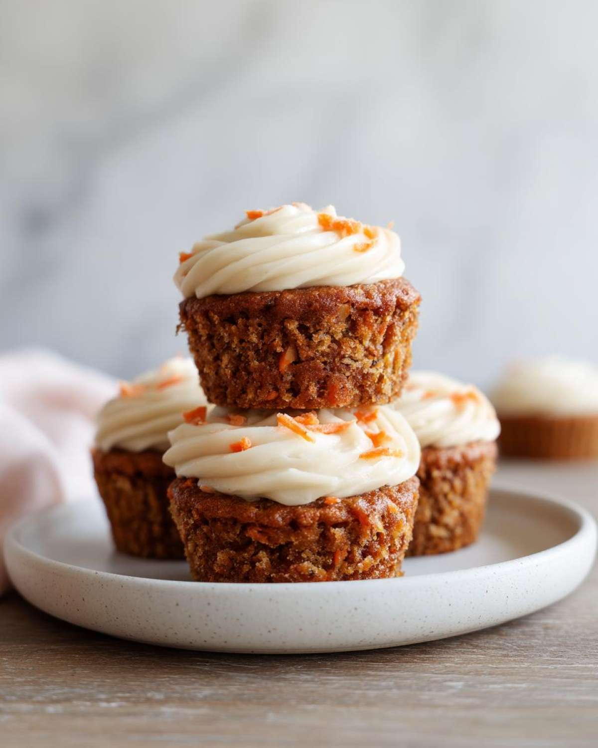 A stack of two Gluten-Free Carrot Cake Cupcakes topped with cream cheese frosting and shredded carrots.