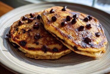 Two fluffy Gluten-Free Chocolate Chip Banana Pancakes stacked on a plate, drizzled with syrup and topped with melted chocolate chips.