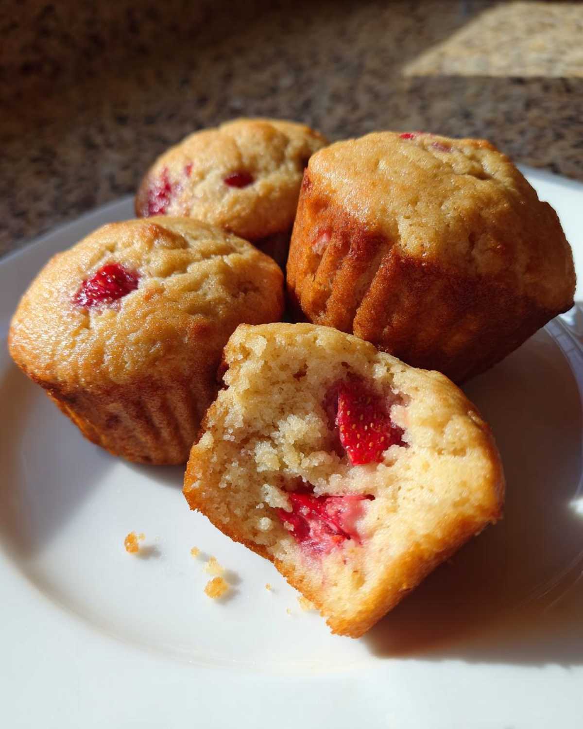 A close-up of several Gluten-Free Strawberry Almond Muffins on a white plate, one broken open showing strawberries inside.