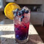Close-up of a tall glass filled with Iced Blueberry Lavender Lemonade, featuring layered purple liquid, ice, fresh blueberries, and a lemon slice.