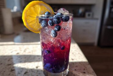 Close-up of a tall glass filled with Iced Blueberry Lavender Lemonade, featuring layered purple liquid, ice, fresh blueberries, and a lemon slice.