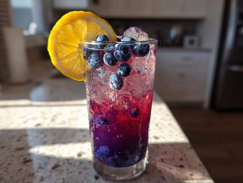 Close-up of a tall glass filled with Iced Blueberry Lavender Lemonade, featuring layered purple liquid, ice, fresh blueberries, and a lemon slice.