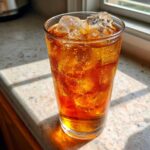 A tall glass filled with cold Iced Peach Green Tea and plenty of ice cubes, sitting on a sunlit counter.