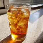 A tall glass filled with amber-colored Iced Peach Green Tea and many ice cubes, sitting in bright sunlight.