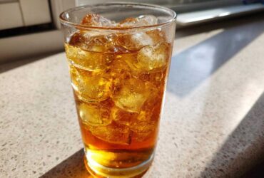 A tall glass filled with amber-colored Iced Peach Green Tea and many ice cubes, sitting in bright sunlight.