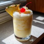 A tall glass filled with a frozen Iced Pineapple Coconut Mocktail, garnished with a pineapple wedge and a maraschino cherry.