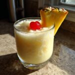 A frosty glass of Iced Pineapple Coconut Mocktail garnished with a maraschino cherry and pineapple wedge.